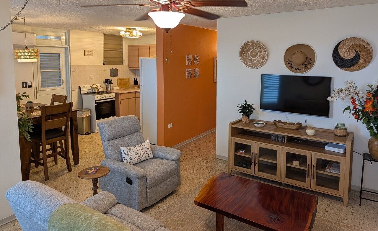 A living room with a sofa, armchair, wooden coffee table, TV, and a kitchen in the background.