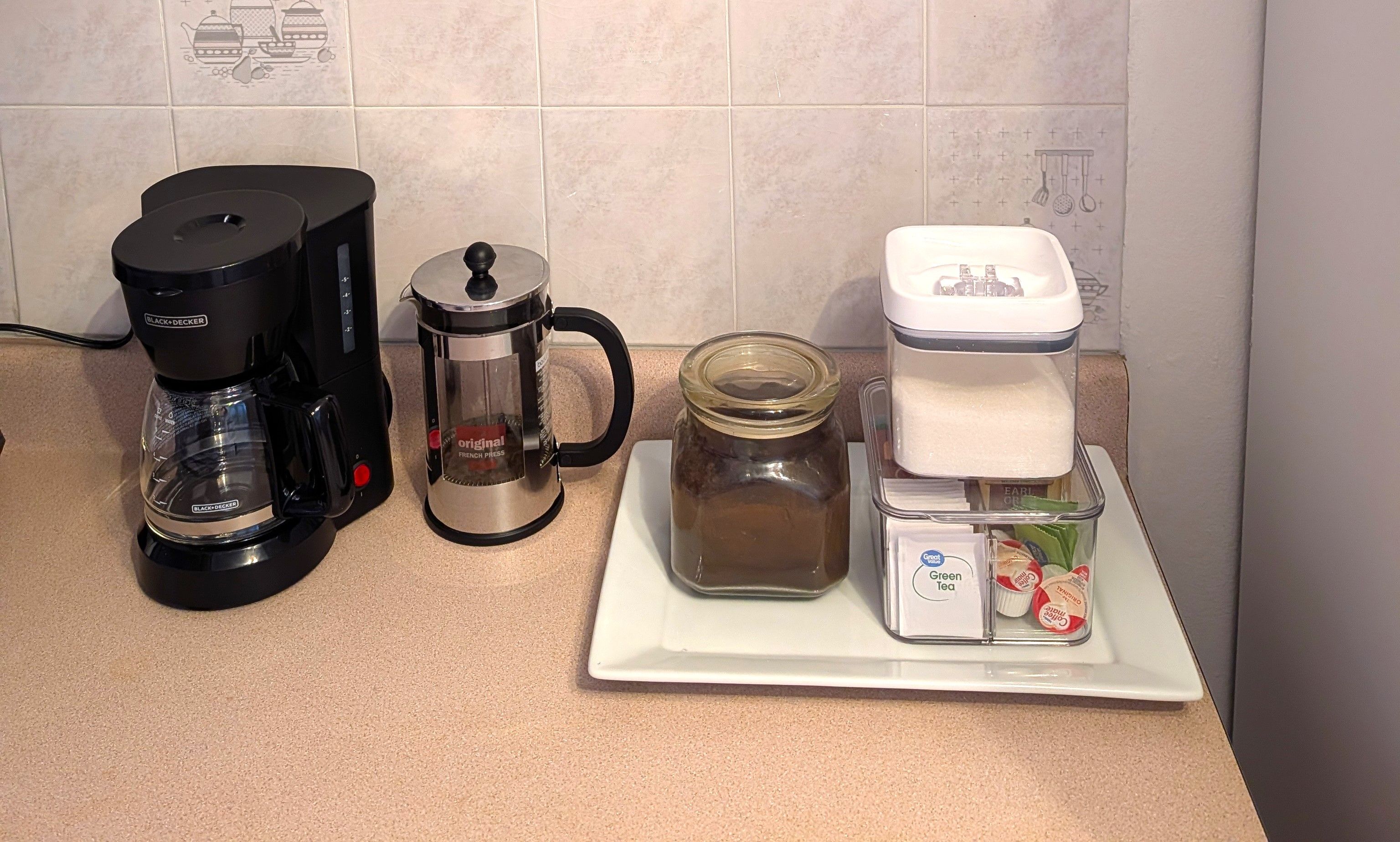En køkkenbordplade med en kaffemaskine, en presskanne, et glas med te og en beholder med grøn te.