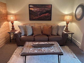 A living room with a brown leather sofa, two side tables with lamps, a framed landscape painting, and a rug. - Walk to Town! River Front Condo-1 King Bedroom-1 Bathroom (Estes Park)