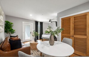 Ein modernes Zimmer mit einem runden Esstisch, einer Couch, einem kleinen Holzschemel und einem großen Fenster mit Vorhängen.