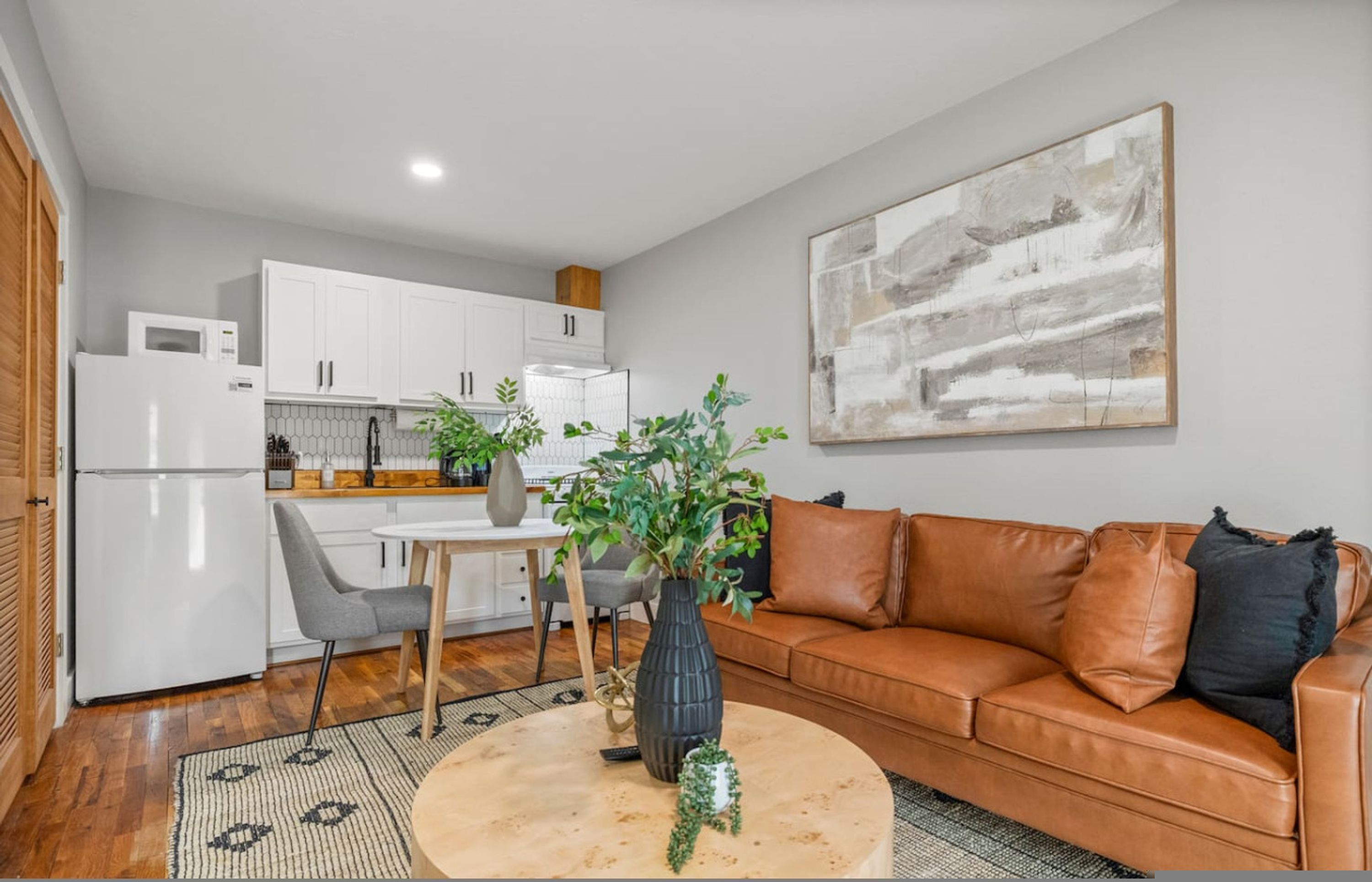 A modern kitchen with white cabinets, a dining table with chairs, a brown leather sofa, and a large abstract painting on the wall.