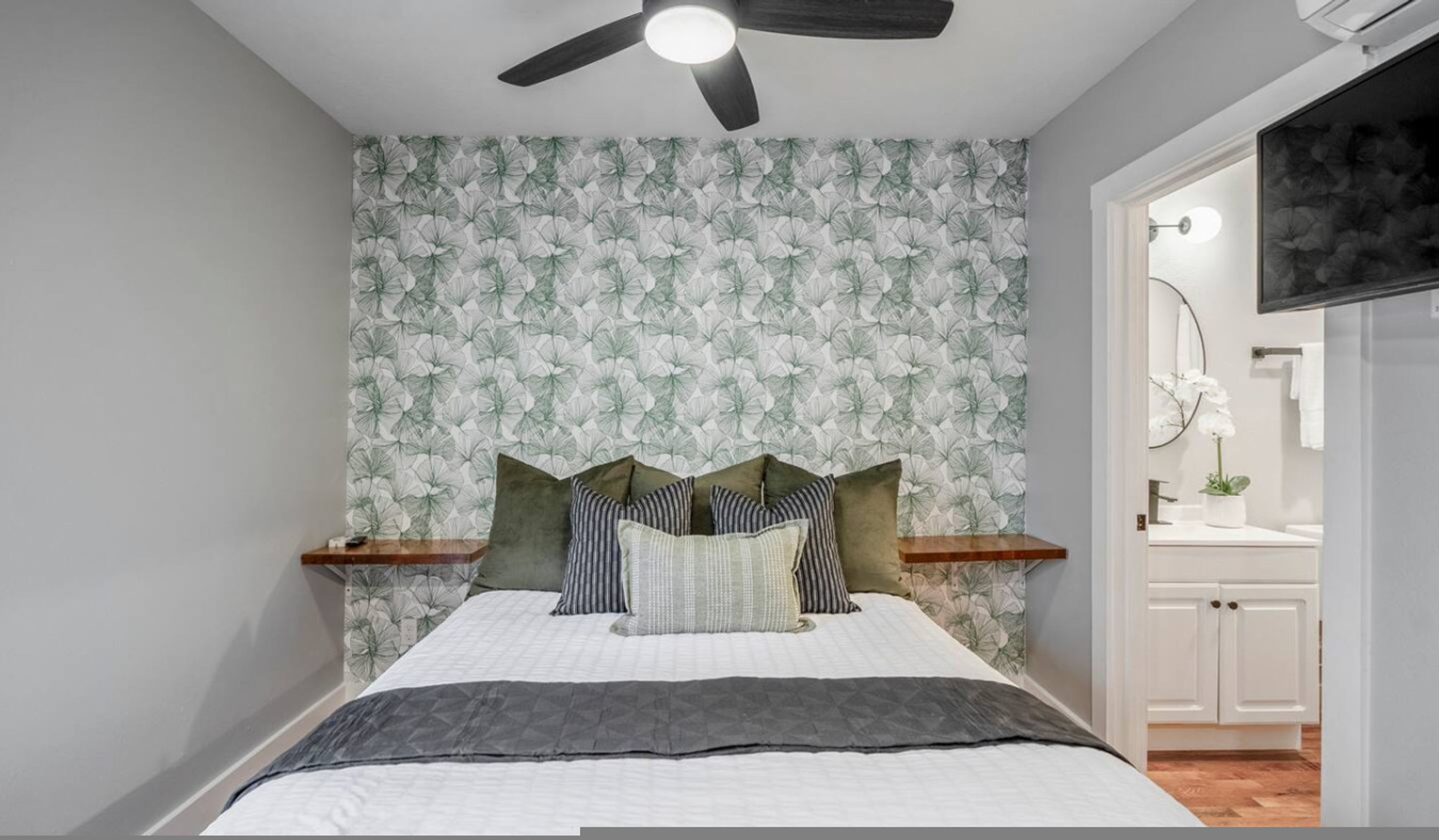Une chambre à coucher avec un lit, un ventilateur de plafond, un mur orné d’un motif de feuilles, et une salle de bain visible à travers une porte ouverte.