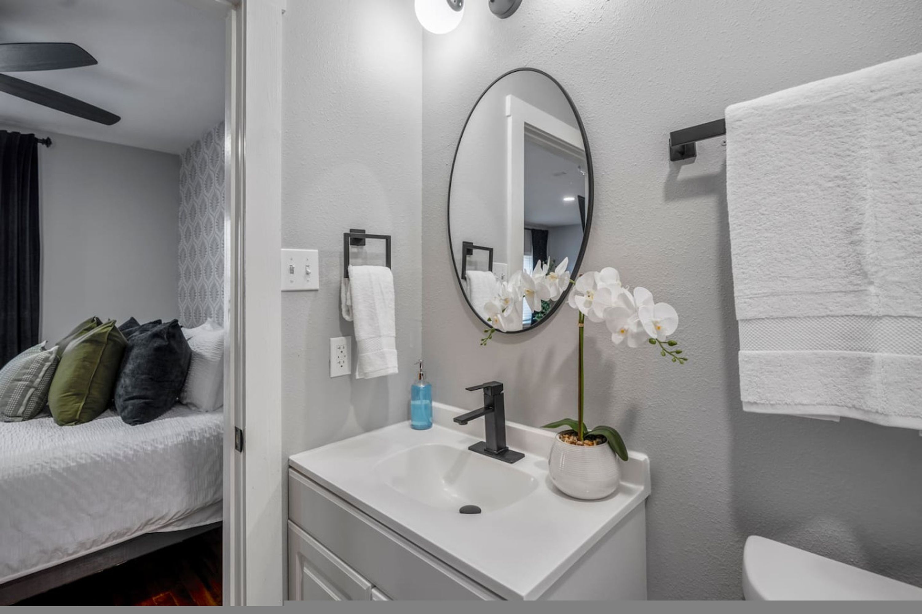 A modern bathroom with a white sink, a mirror, a towel rack, and a bed with pillows.