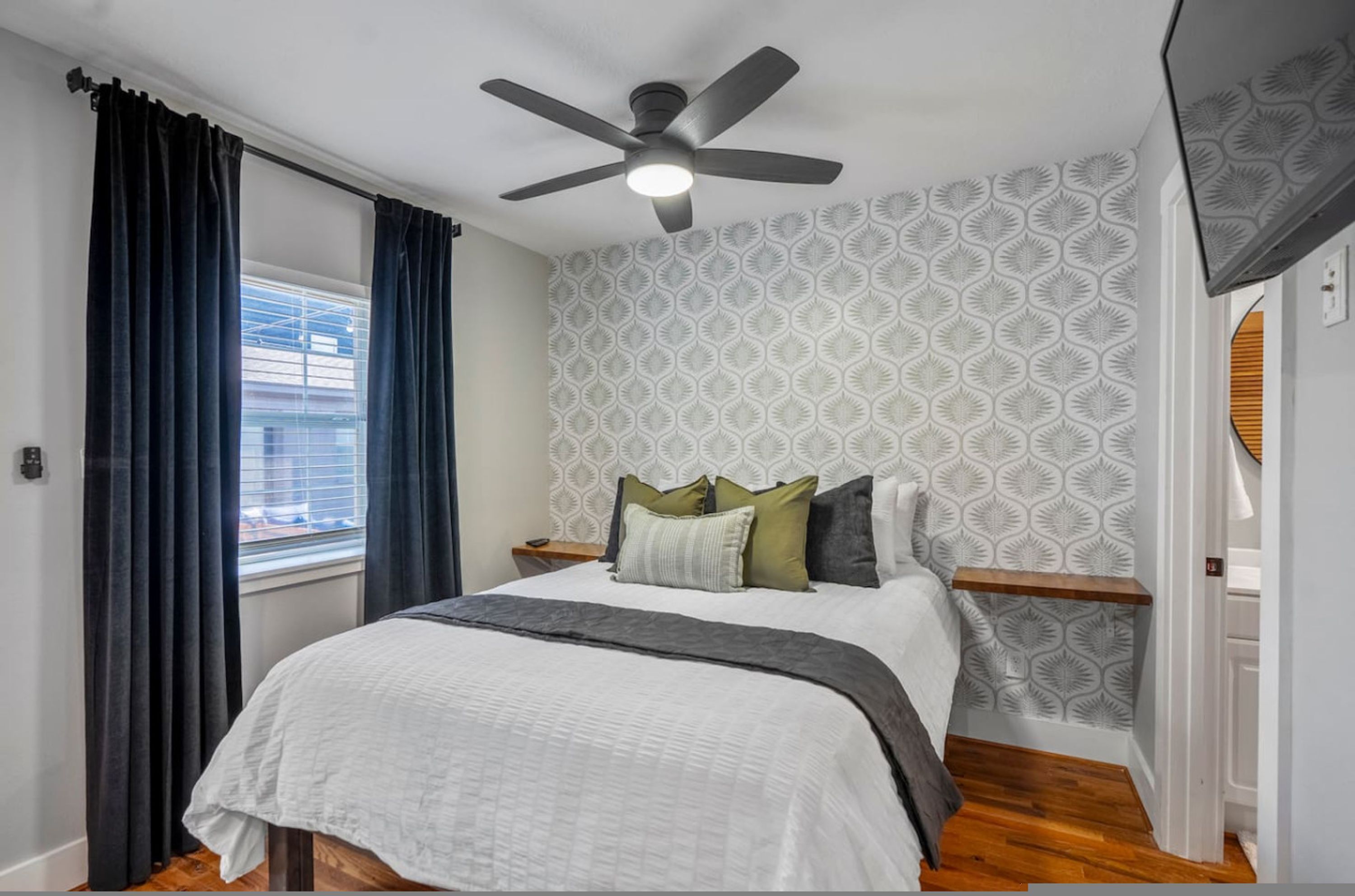 A bedroom with a bed, a ceiling fan, a window with curtains, and patterned wallpaper.