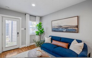 Ein Wohnzimmer mit einem blauen Sofa, einer weißen Tür, einem Fenster mit Vorhängen und einem Gemälde an der Wand.
