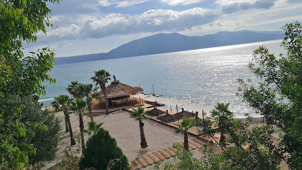 Ein Strand mit Palmen, einem mit Stroh gedeckten Gebäude und Blick auf das Meer und die Berge.