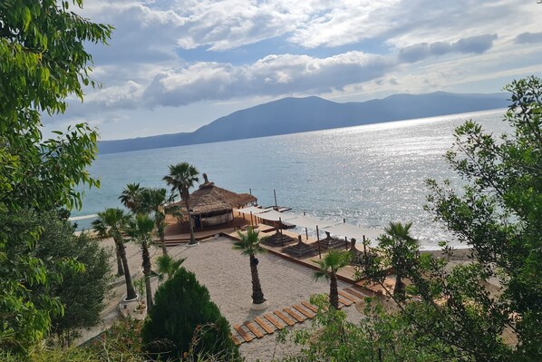Ein Strand mit Palmen, einem mit Stroh gedeckten Gebäude und Blick auf das Meer und die Berge.