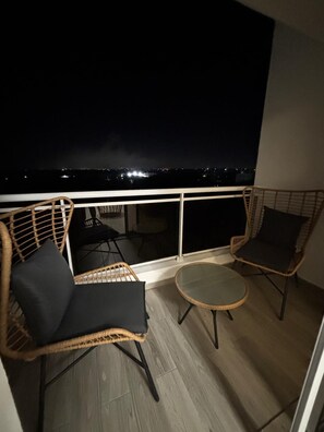 Ein Balkon mit Korbstühlen und einem kleinen Tisch, mit Blick auf eine nächtliche Stadtlandschaft.