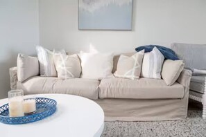 A beige sofa with multiple pillows, a white coffee table with a blue tray, and a framed picture on the wall. - Bay Breeze Retreat, Glenelg, Absolute Beachfront (Glenelg)