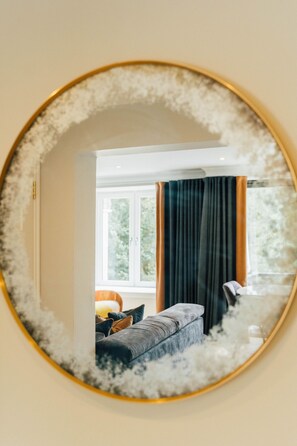 Ein runder Spiegel mit goldener Rahmung, der einen Raum mit Fenster, Vorhängen und einer Couch reflektiert.