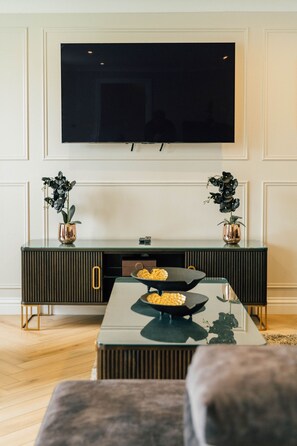 A living room with a flat-screen TV mounted on the wall, a dark-colored entertainment unit with two potted plants, a glass coffee table with snacks, and a plush sofa. - Eagleside Apartments - 21 - sleeps 4 guests  in 2 bedrooms (Auchterarder)