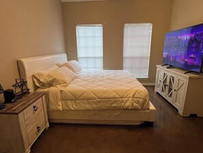 Ein Schlafzimmer mit einem Bett, einer Kommode, einem Fernseher und zwei Fenstern mit Jalousien.