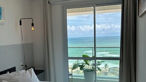 A hotel room with a bed, a bedside lamp, a view of the ocean, and a balcony with a plant. - 180-degree sea view (Salvador)