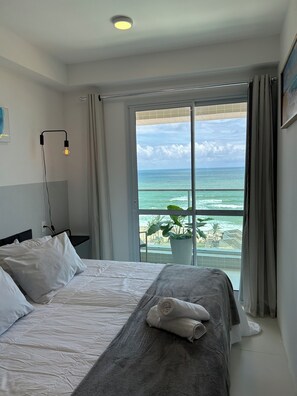 A hotel room with a bed, a bedside lamp, a view of the ocean, and a balcony with a plant. - 180-degree sea view (Salvador)