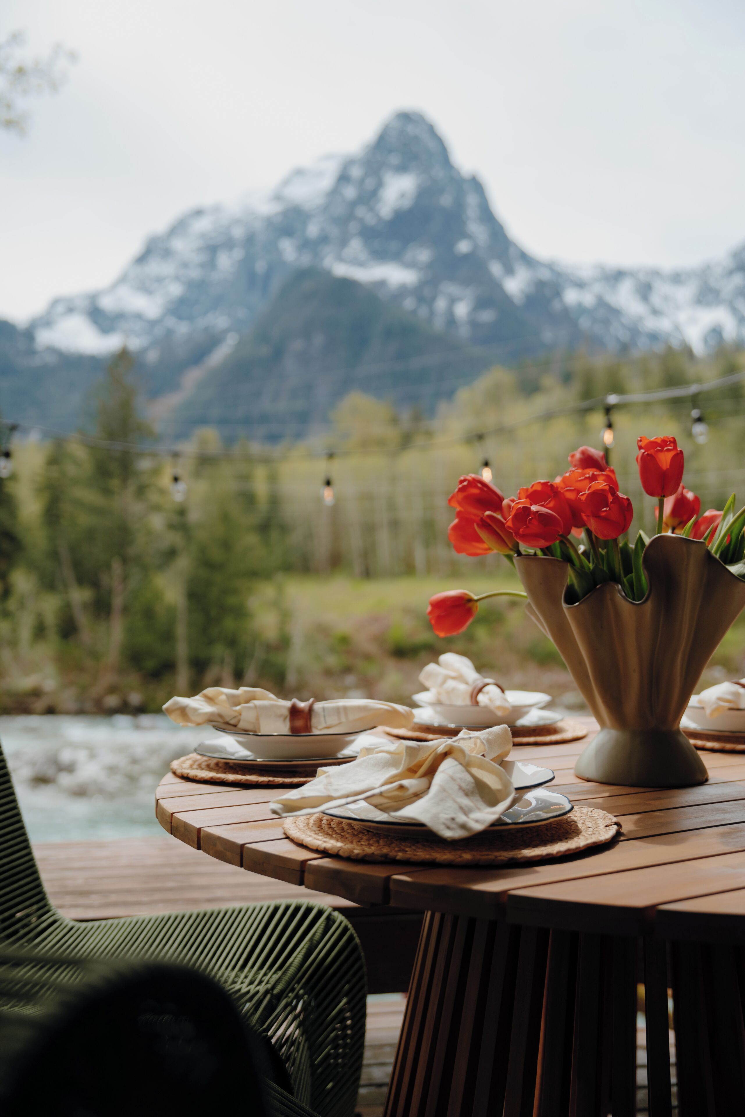 A table with a vase of red tulips, plates, and napkins on a wooden deck with a mountain view in the background.