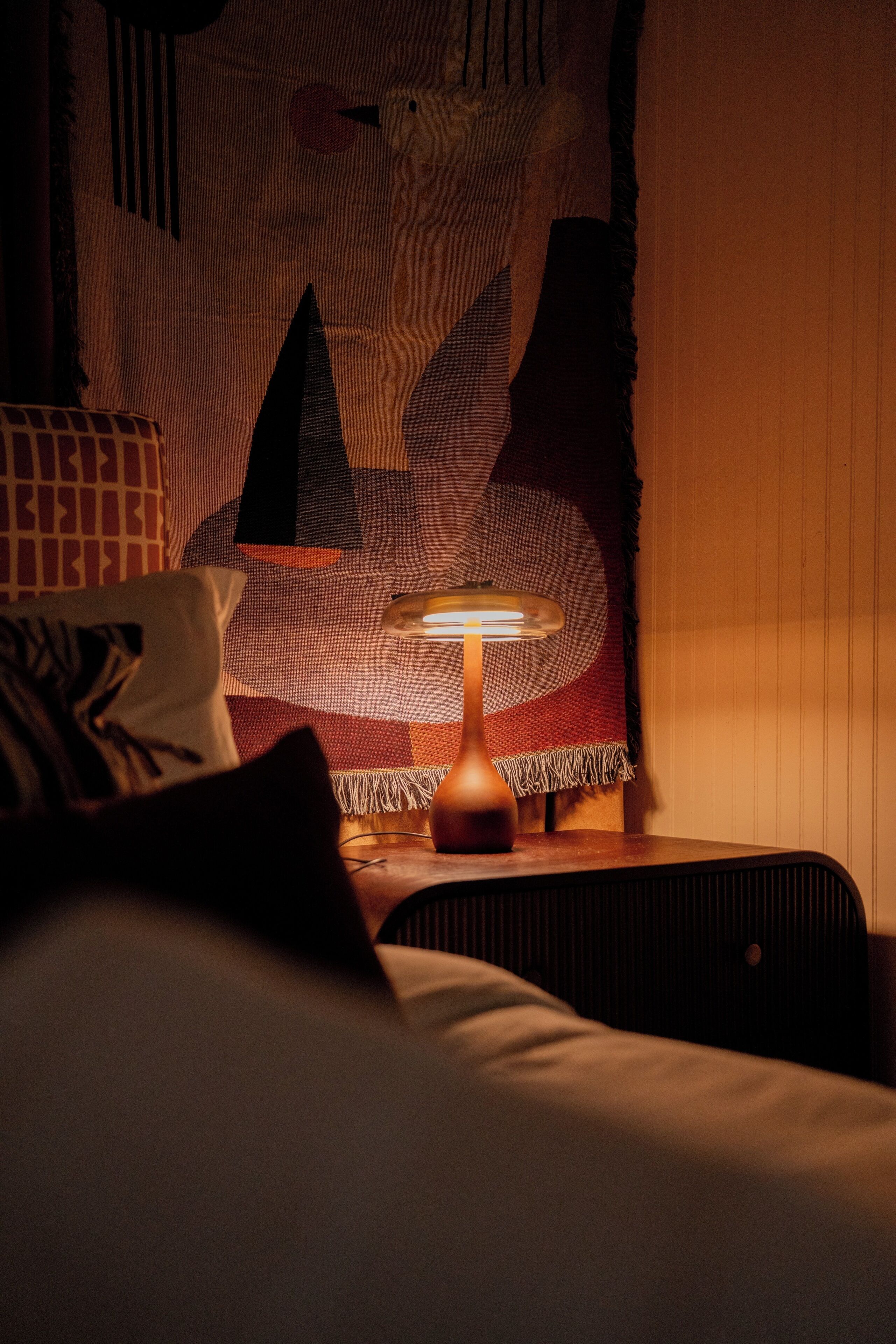 A dimly lit room with a bed, a lamp, and a wall tapestry.