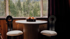 A room with a round table, two chairs, a window with a view of mountains and trees, and a wooden floor.