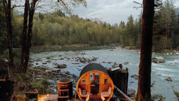 A wooden cabin by a river with a fireplace, seating area, and a view of the surrounding forest.