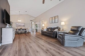 A spacious living room with a wooden floor, a leather sofa, a leather armchair, a TV mounted on the wall, and a dining area with a table and chairs. - Gorgeous 3BR/2BA Saltwater Pool on Rivard Public Golf Course (Brooksville)