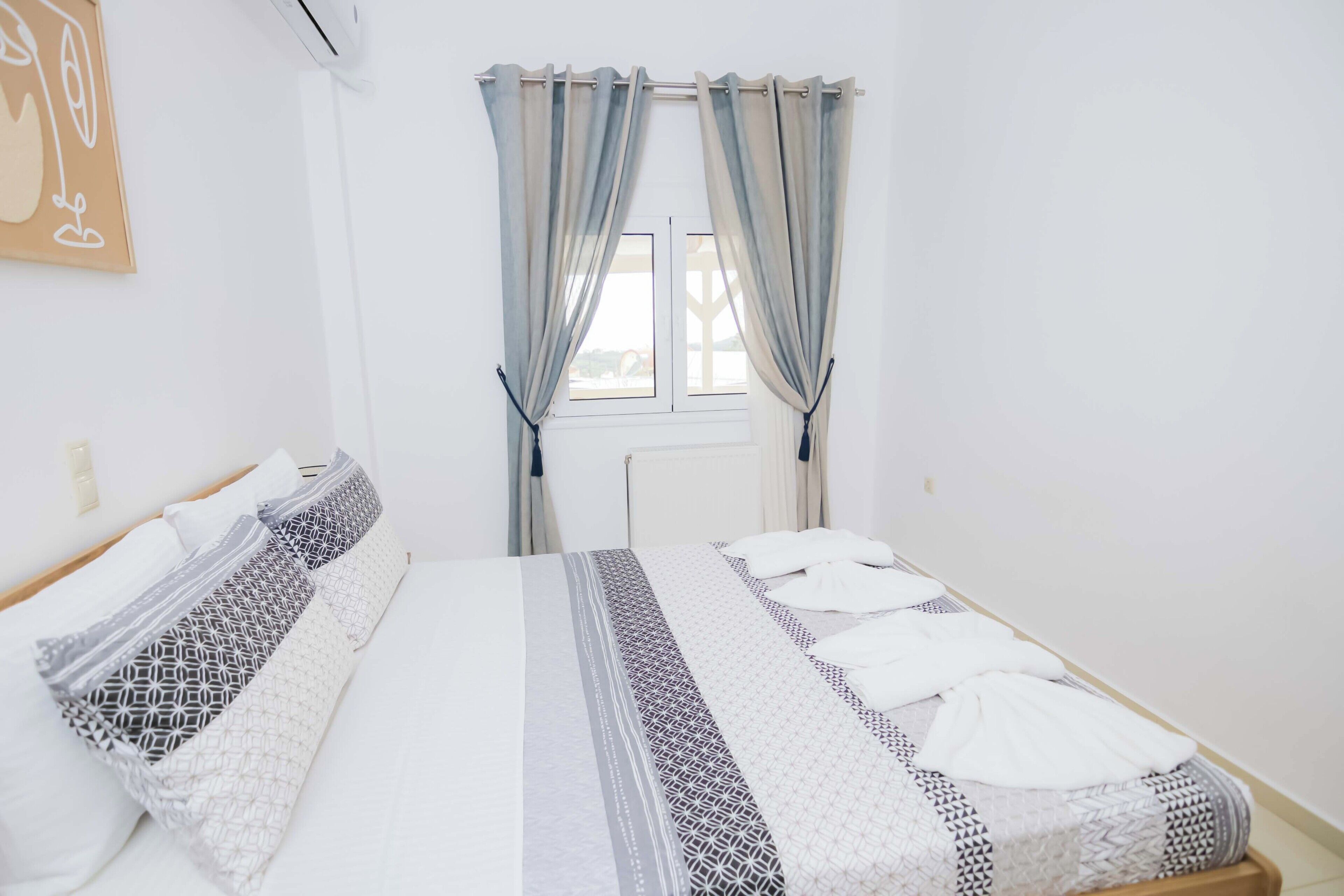 Ein sauberes, helles Hotelzimmer mit einem ordentlich bezogenen Bett, einem Fenster mit Vorhängen und einem an der Wand hängenden Kunstwerk.