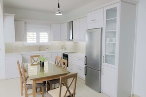 A modern kitchen with white cabinets, a stainless steel refrigerator, and a dining table with chairs. - Thea Villa (Faistos)
