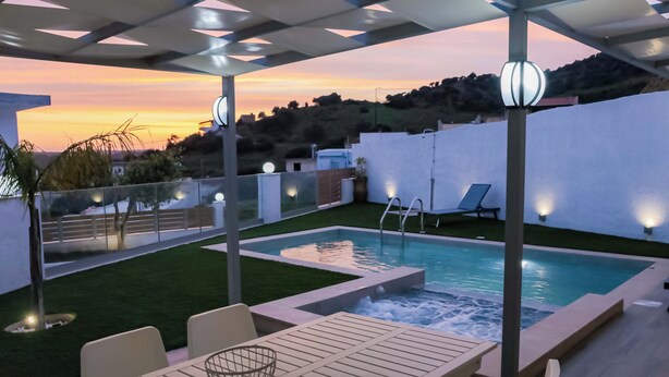 A modern outdoor pool area with a table and chairs, surrounded by a well-maintained lawn and a view of the sunset. - Thea Villa (Faistos)