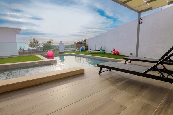 A modern outdoor pool area with a wooden deck, lounge chairs, and a slide. - Thea Villa (Faistos)