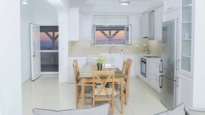 A modern kitchen with white cabinets, a dining table with chairs, a refrigerator, and a window with a view. - Thea Villa (Faistos)