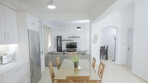 A modern kitchen with white cabinets, a stainless steel refrigerator, and a dining area with a wooden table and chairs. - Thea Villa (Faistos)
