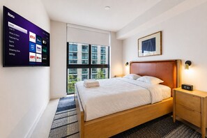 A hotel room with a bed, a nightstand, a wall-mounted TV displaying streaming services, a large window, and a framed picture on the wall. - Corner 2BR with Skyline Views and Rooftop Pool Minutes to South Beach, Brickell and Wynwood (Miami)