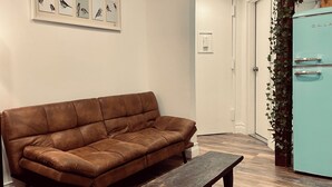 A brown leather sofa, a wooden bench, and a teal refrigerator in a room with wooden flooring and a white door. - Quiet LES Designer Retro-Chic 1 Bedroom Apt Near Everything (New York)