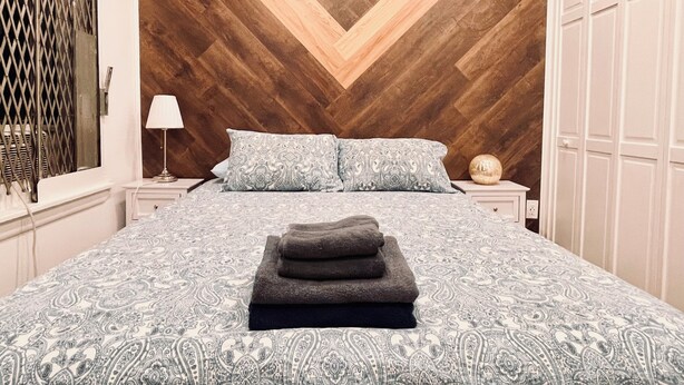 A bedroom with a herringbone-patterned wooden ceiling, a patterned bedspread, two pillows, a bedside lamp, a round decorative object, and a stack of folded towels. - Quiet LES Designer Retro-Chic 1 Bedroom Apt Near Everything (New York)