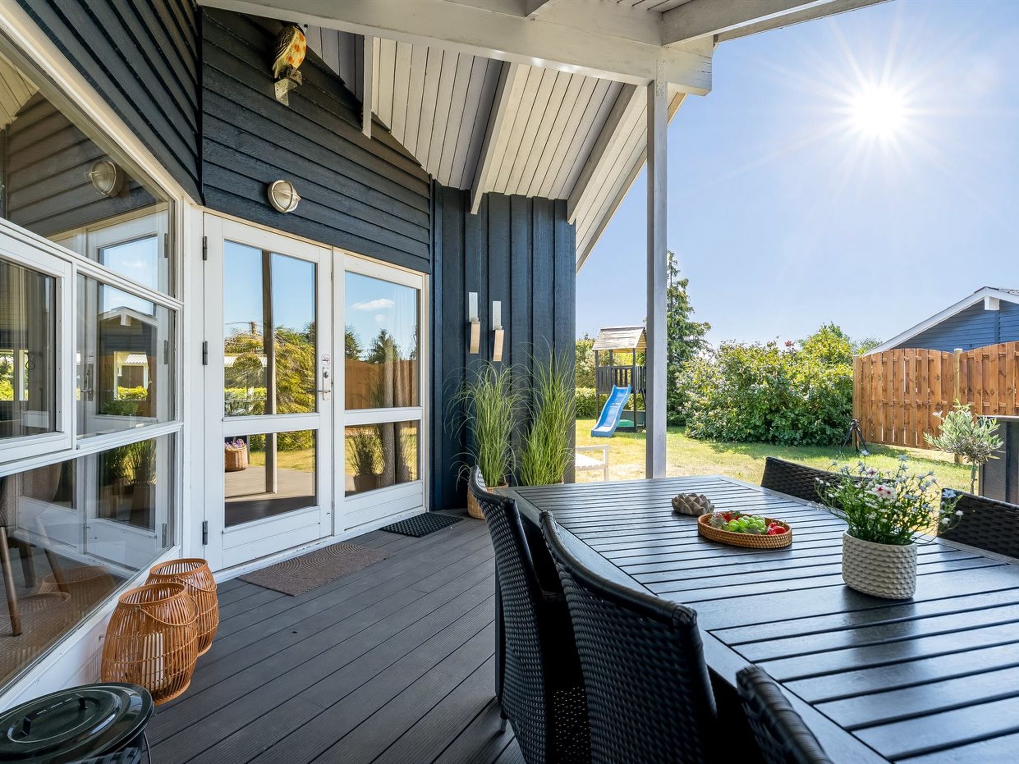 En overdækket terrasse med et bord og stole, en skydedør og udsigt til en baghave med en rutchebane.
