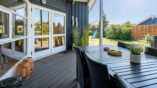 Eine überdachte Terrasse mit Tisch und Stühlen, eine Glastür und Blick auf einen Garten mit einer Rutsche.
