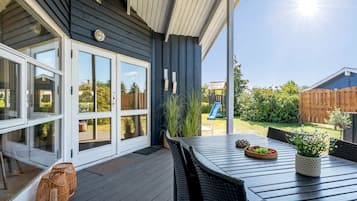 En overdækket terrasse med et bord og stole, en skydedør og udsigt til en baghave med en rutchebane.