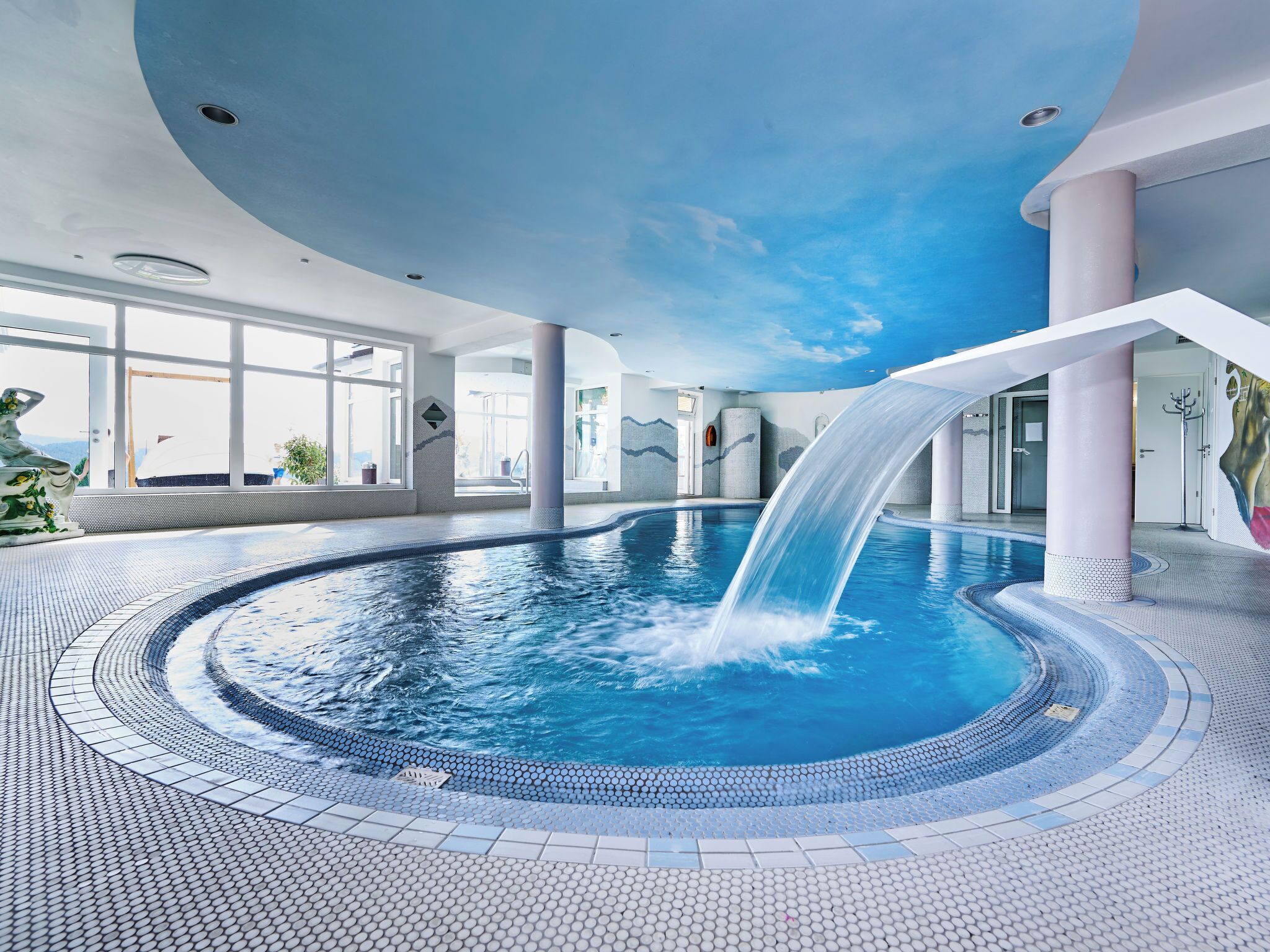 Una gran piscina interior amb cascada, envoltada per un paviment de rajoles i grans finestres.