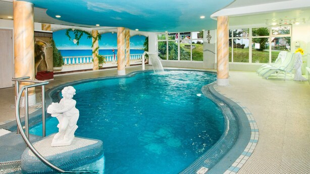 An indoor swimming pool with a marble statue, a waterfall feature, and a view of the outdoors through large windows. - Heckenrose mit Badewelt und Sauna inkl. by Interhome (Sankt Englmar)