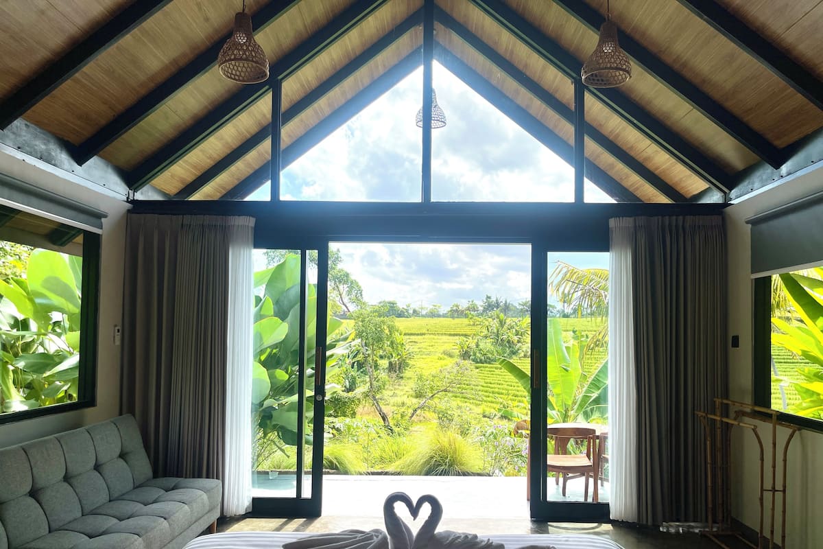 Quarto com cama, sofá e vista para uma paisagem exuberante através de grandes janelas.