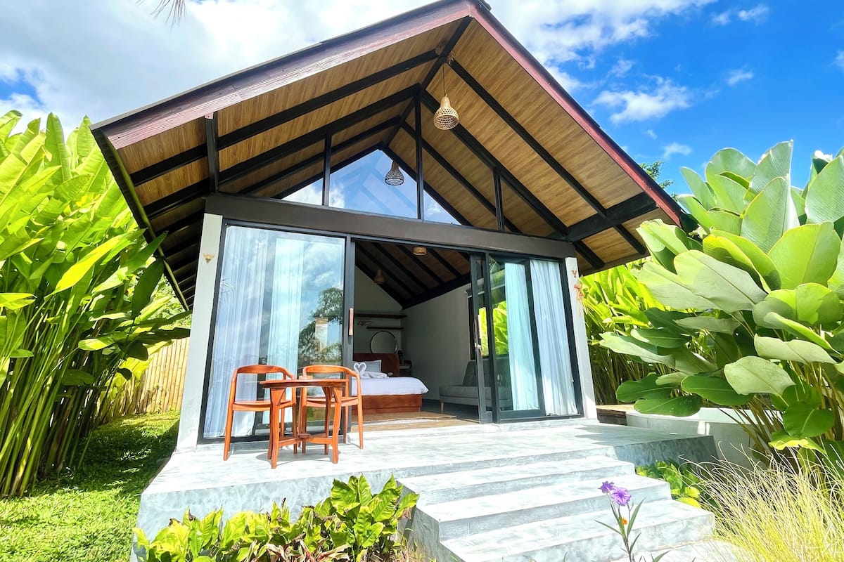 Uma casa moderna com uma grande porta de vidro, um deck de madeira e uma cama visível no interior.