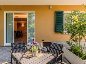 An outdoor patio with a table set for two, a chair, a potted plant, and a view of the interior through a glass door. - Apartment Renza by Interhome (Colico)