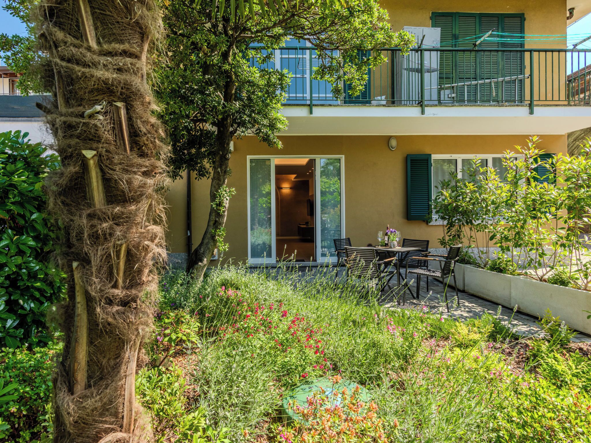 A two-story building with a balcony, a garden with various plants and flowers, and a dining area with outdoor furniture.
