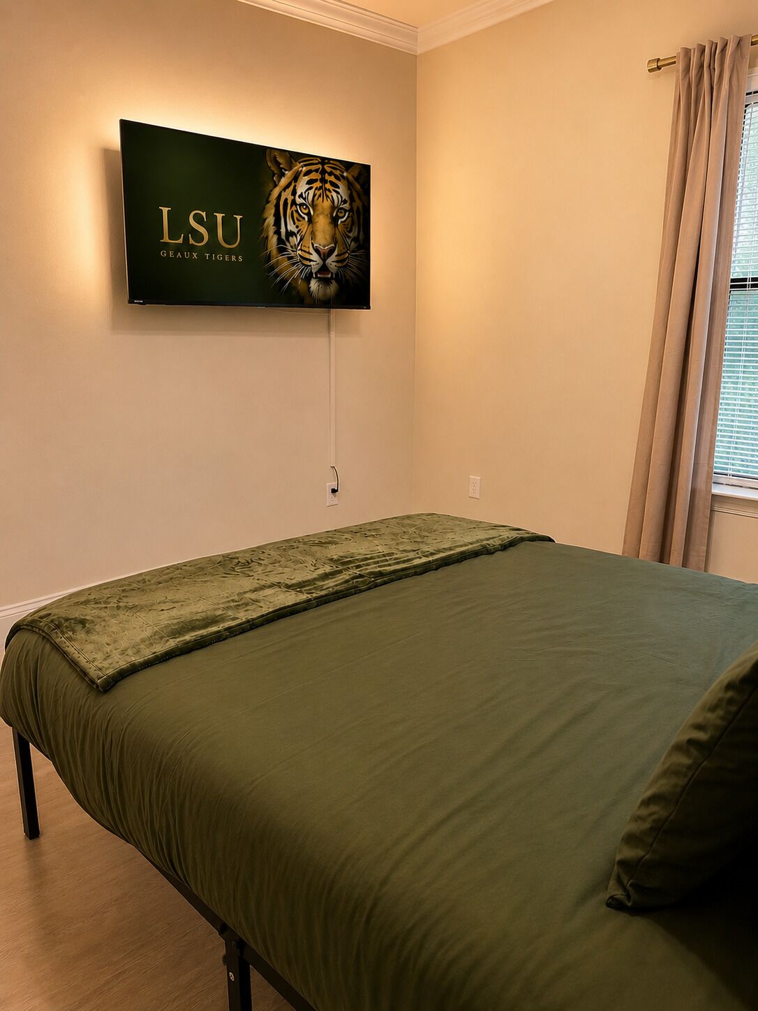 A bedroom with a bed, a wall-mounted TV displaying LSU Gators, and a window with curtains.