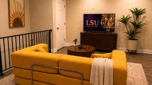 En stue med en gul sofa, et fjernsyn der viser LSU Gators, en potteplante og en dekorativ vægdekoration.