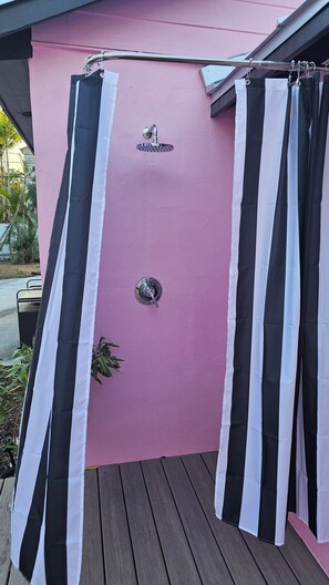 Un'area doccia esterna rosa con tende a righe bianche e nere, un soffione metallico e una piccola pianta.