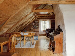 Ein gemütliches Dachzimmer mit Holzbalken, einem Klavier, einem Kamin, einem Bücherregal und einem Essbereich.