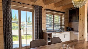 A wooden interior with a dining table, chairs, a kitchen area, and a view through the window.