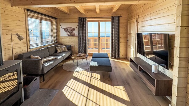 A wooden cabin interior with a sofa, TV, and a large window overlooking a deck.
