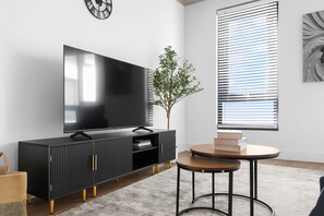 Ein modernes Wohnzimmer mit Flachbildfernseher, einer schwarzen Unterhaltungsanlage, einem runden Couchtisch und einem großen Fenster mit Jalousien.
