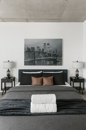 Une chambre d’hôtel moderne avec un grand lit, deux tables de chevet, un tableau représentant un paysage urbain et une vue sur un pont.