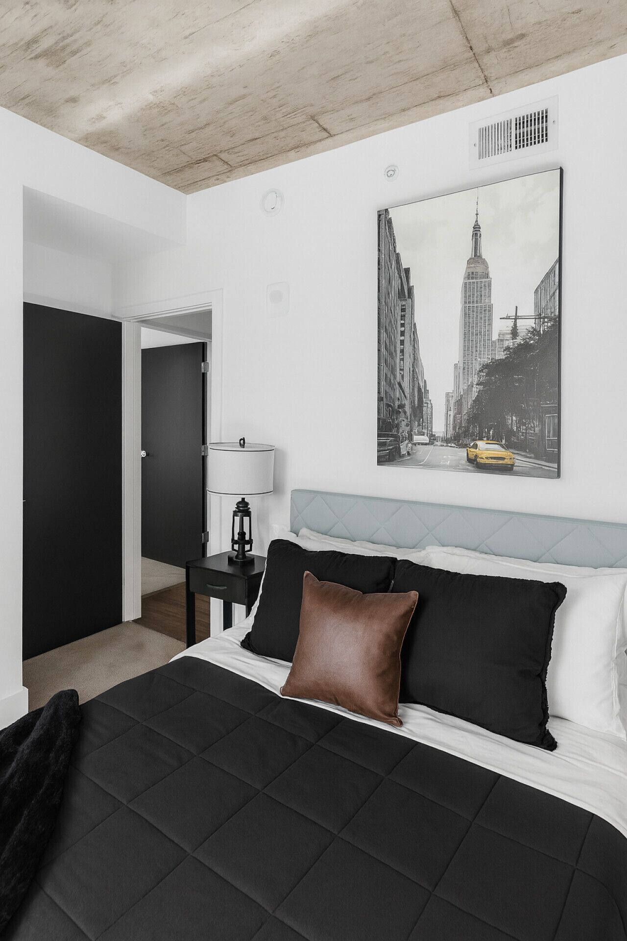 A bedroom with a bed, a nightstand, a lamp, a door, and a cityscape painting on the wall.